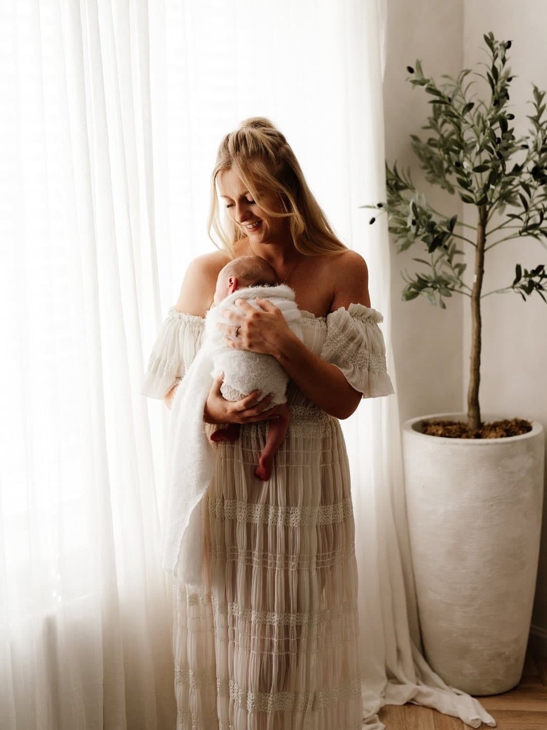 Smiling blonde woman in a white dress cradles a swaddled newborn baby near a window.