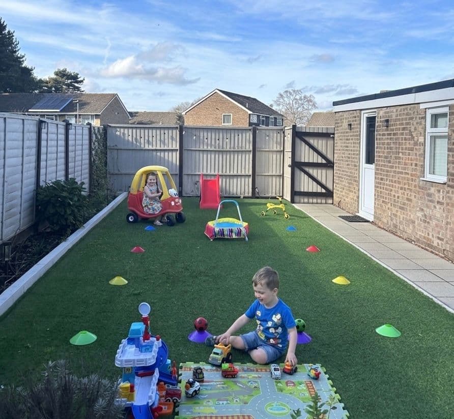 Two children playing with colorful toys on an artificial lawn in a fenced backyard.