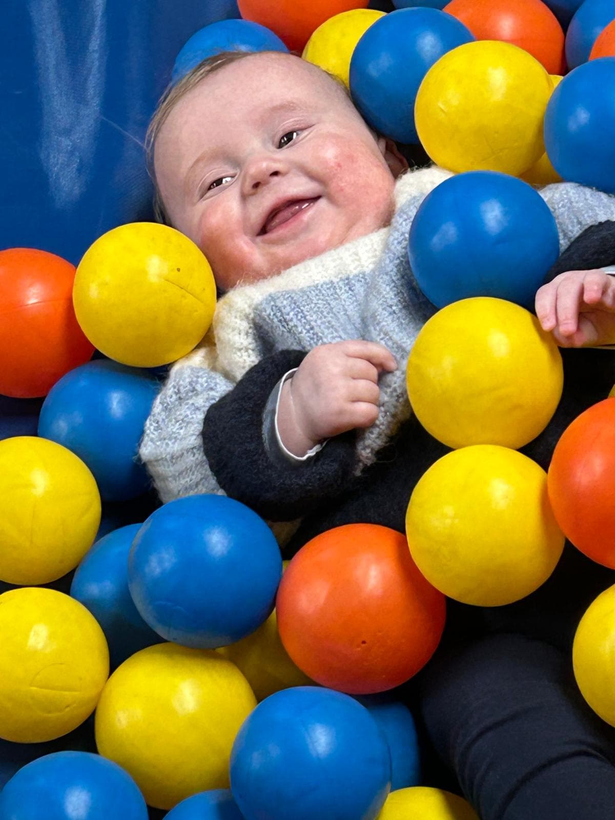 Smiling baby in a sweater lies among blue, yellow, and orange ball pit balls.