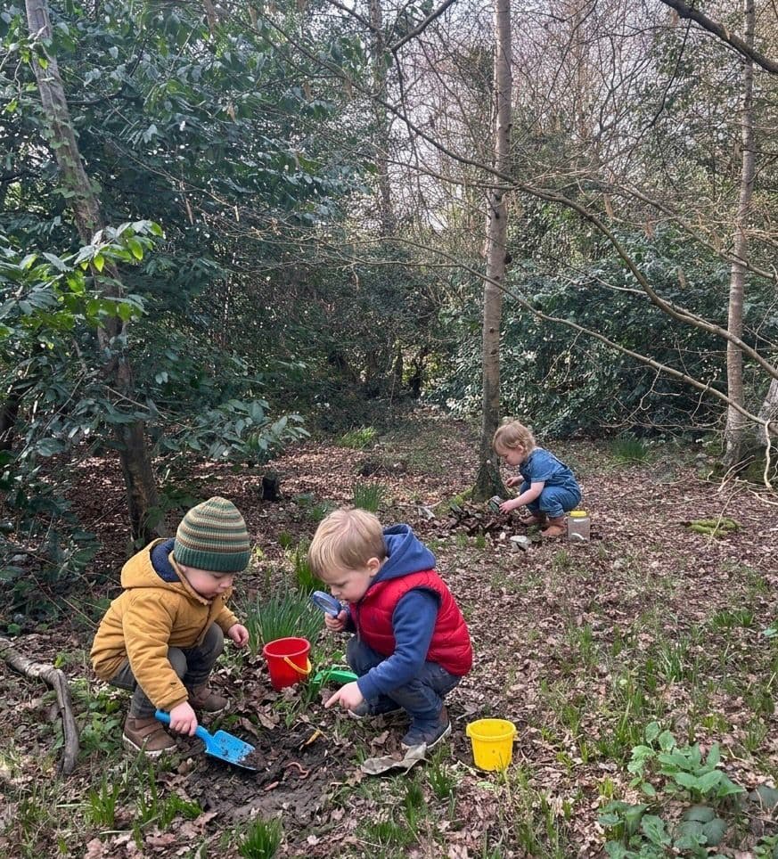 Three young children explore a wooded area with colorful buckets, shovels, and a magnifying glass.